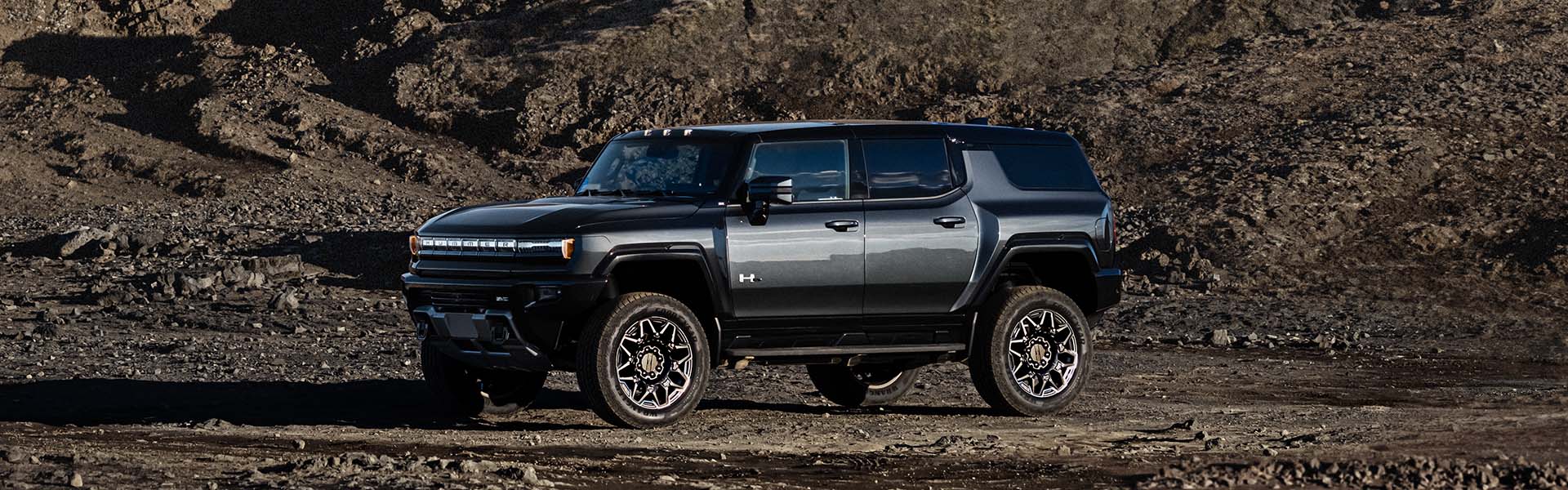 2025 GMC Hummer EV SUV in dark gray parked on rugged off-road terrain, highlighting its electric powertrain, and bold design.
