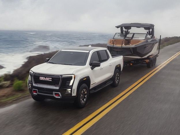 White 2026 GMC Sierra EV towing a boat on a trailer along a coastal highway, with ocean waves and foggy skies in the background.
