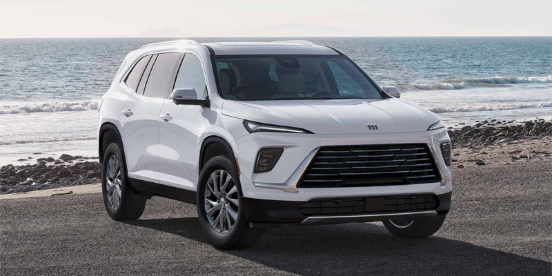 A white Buick Enclave is parked on a coastal road with the ocean in the background. The car's sleek design and chrome details convey a modern, adventurous feel.