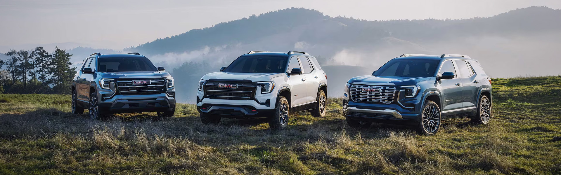Three 2026 GMC Terrain SUVs parked on grassy terrain with mountains in the background.