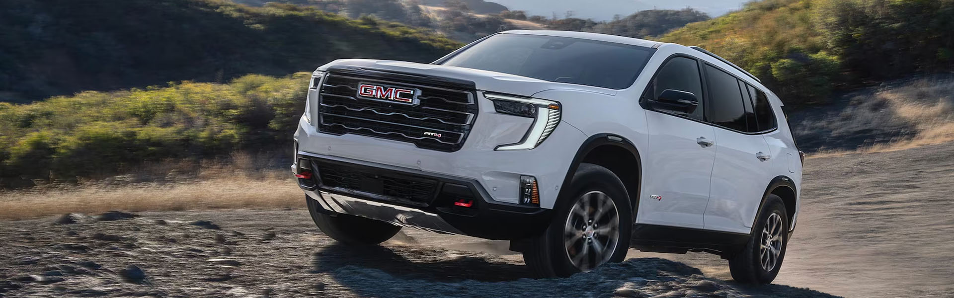 2026 GMC Acadia midsize SUV driving on a rugged dirt trail with a mountain backdrop.
